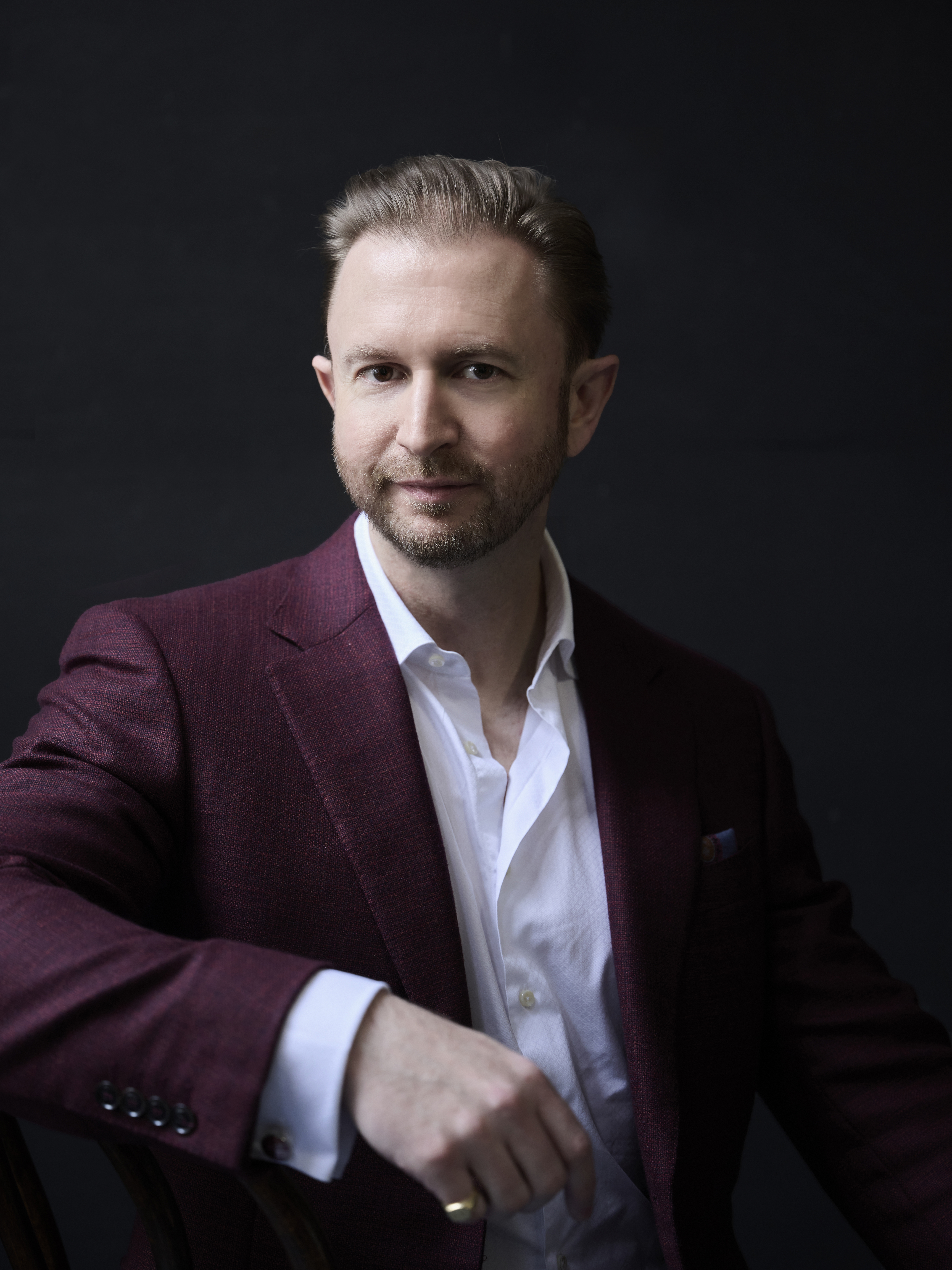 A person with a well-groomed beard and short hair, wearing a maroon blazer and white shirt, sitting against a dark background.