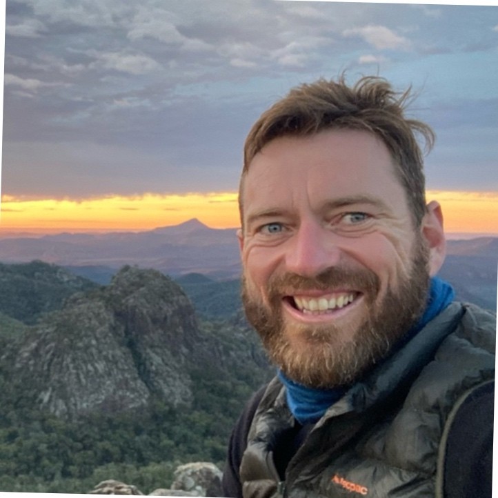 A smiling person with a beard and short hair, wearing a vest and scarf, poses in front of a scenic sunset view with mountains in the background.